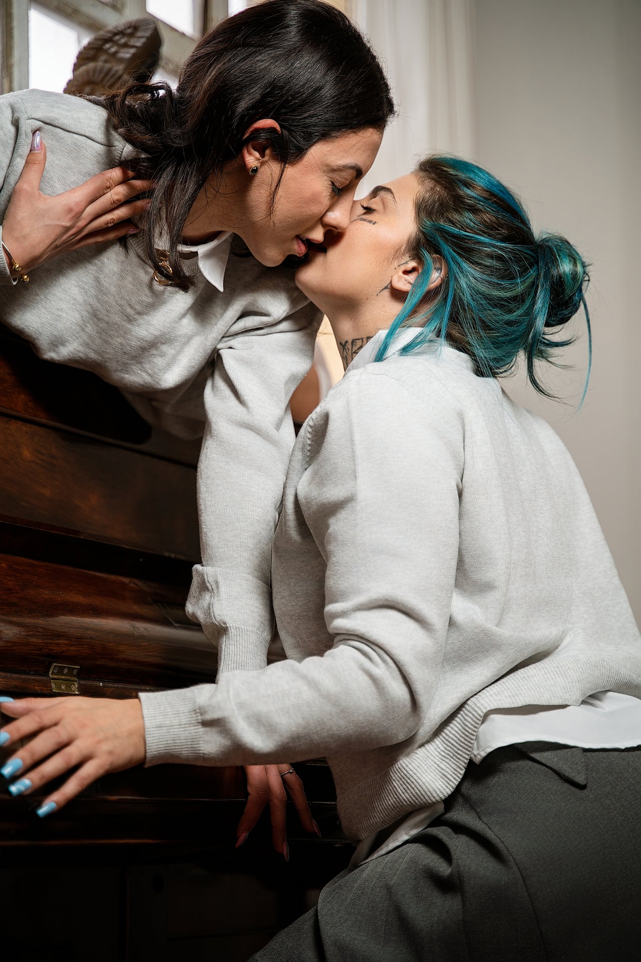 Dues dones amb jerseis de punt clars s'inclinen i es fan un petó vora un piano vertical de fusta al costat d'una finestra alta i lluminosa; una té els cabells foscos i l'altra blau turquesa.