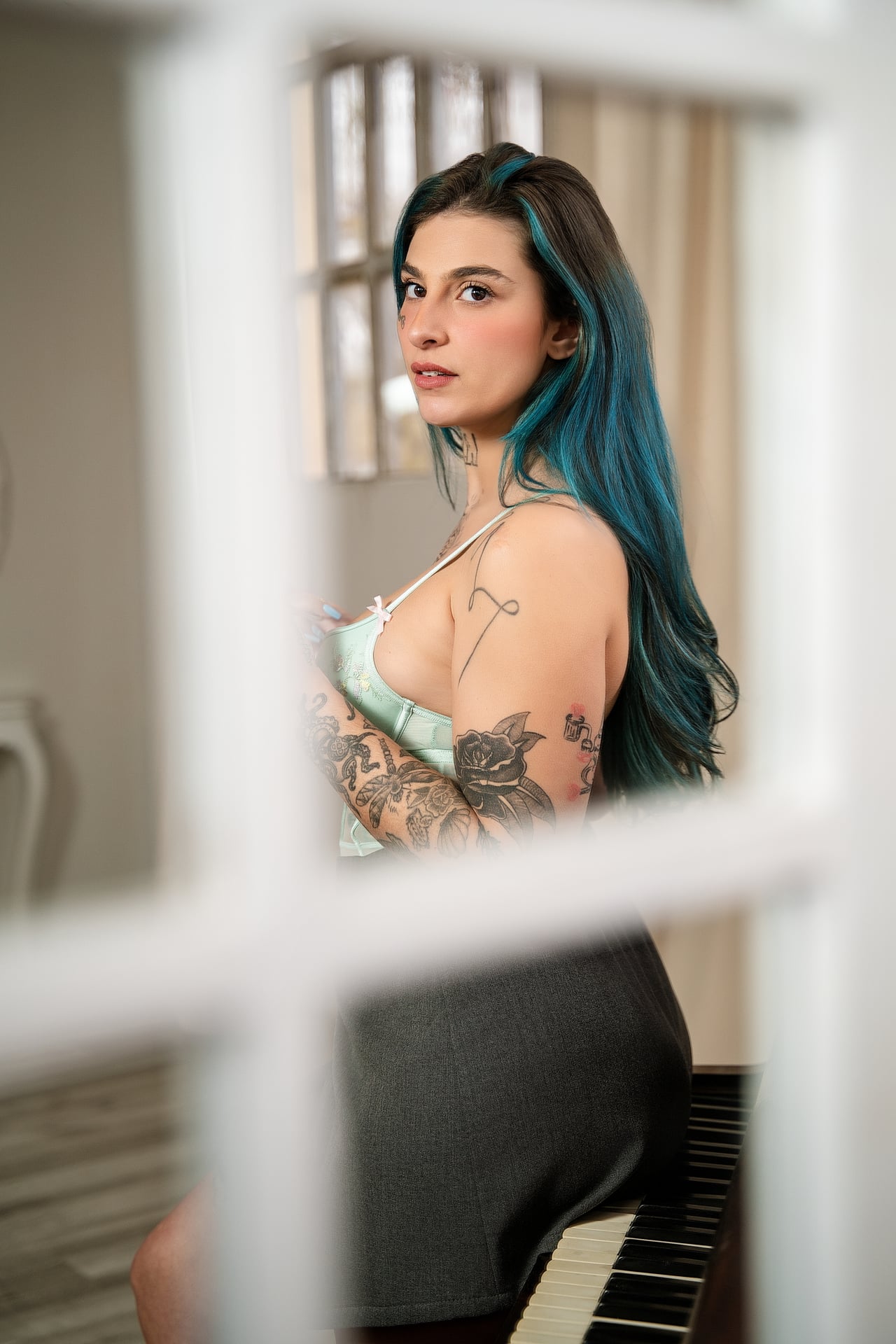 Dona de cabells blaus i braços tatuats, amb top de blonda verd menta i faldilla grisa, dreta de costat al costat d'un piano, vista a través d'un marc de finestra blanc.