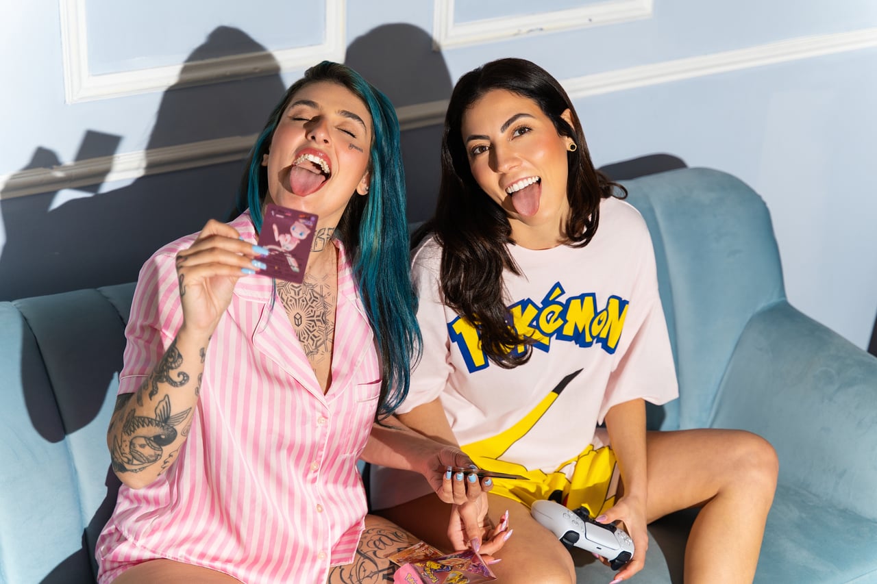 Dues models assegudes en un sofà de vellut blau, una amb els cabells blaus i pijama de ratlles roses sostenint una carta i l'altra amb samarreta ampla de Pokémon i comandament a les mans, totes dues somrient amb la llengua fora.