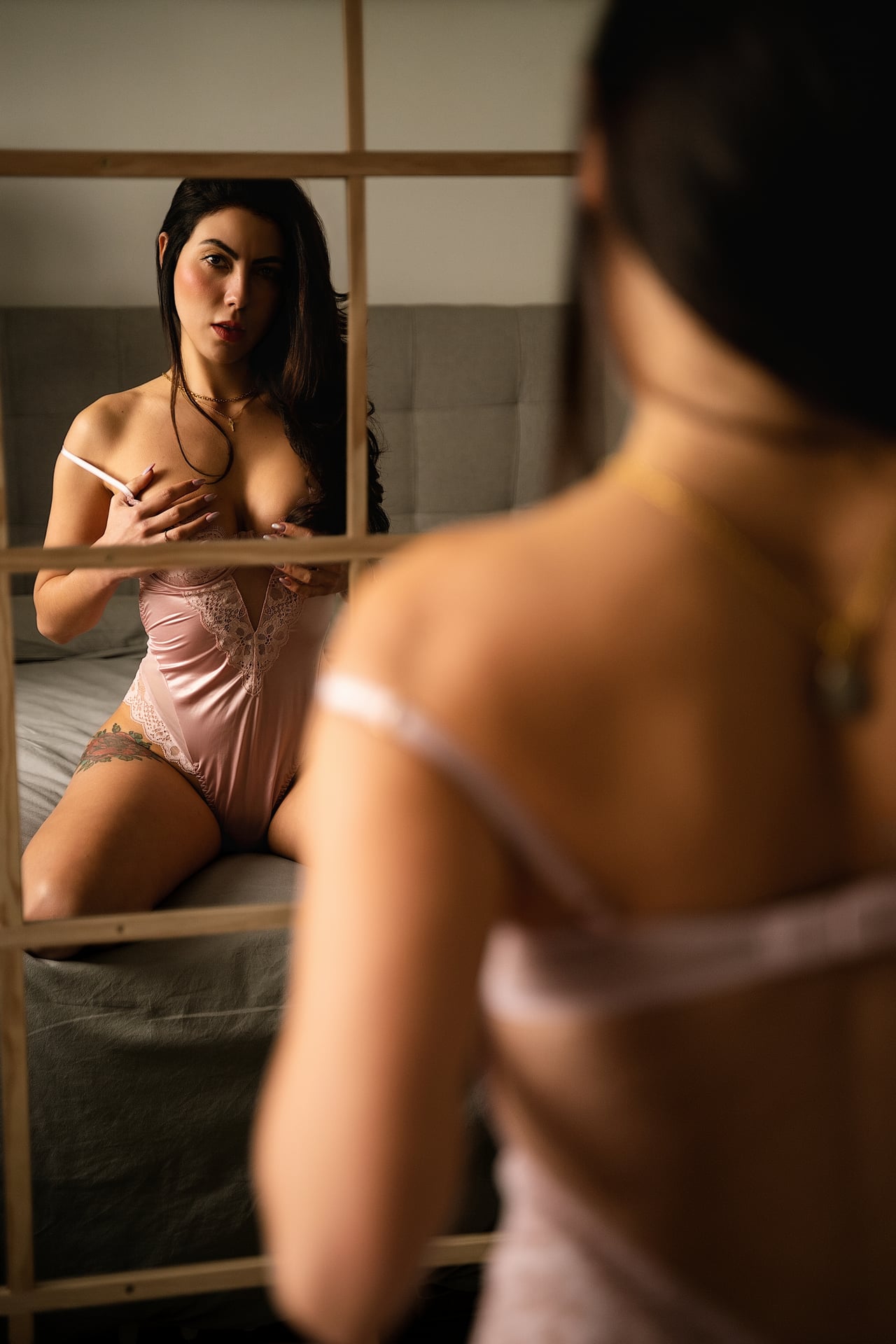 Dona amb lligeria de randa rosa asseguda al llit, reflectida en un mirall amb travessers de fusta; en primer pla, l'esquena nua del seu reflex.