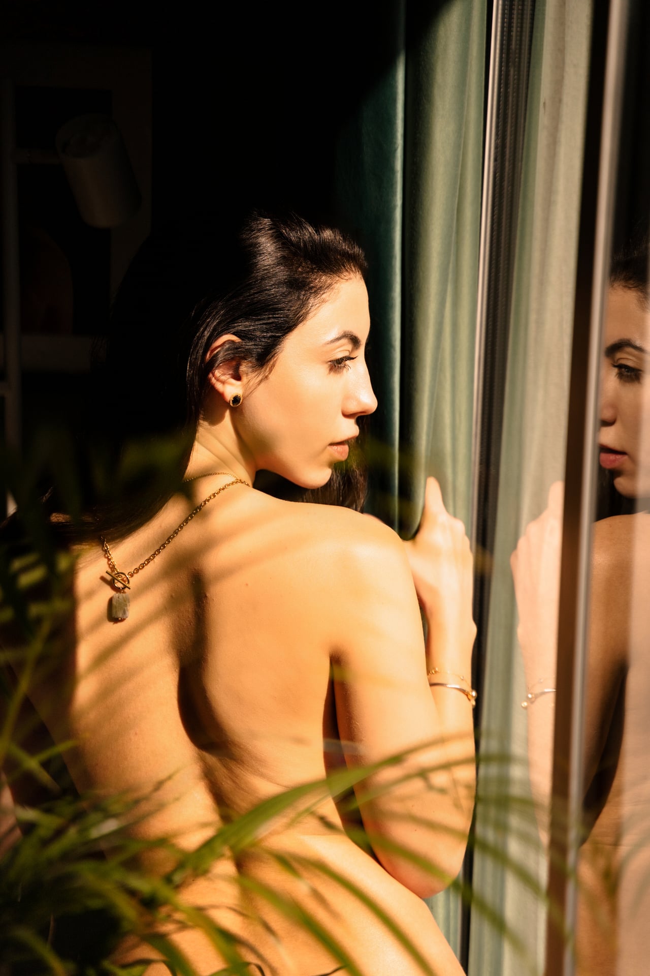 La Gabi, d'esquena i amb l'esquena nua, mira per la finestra mentre el seu reflex apareix al vidre, il·luminada per llum natural càlida.