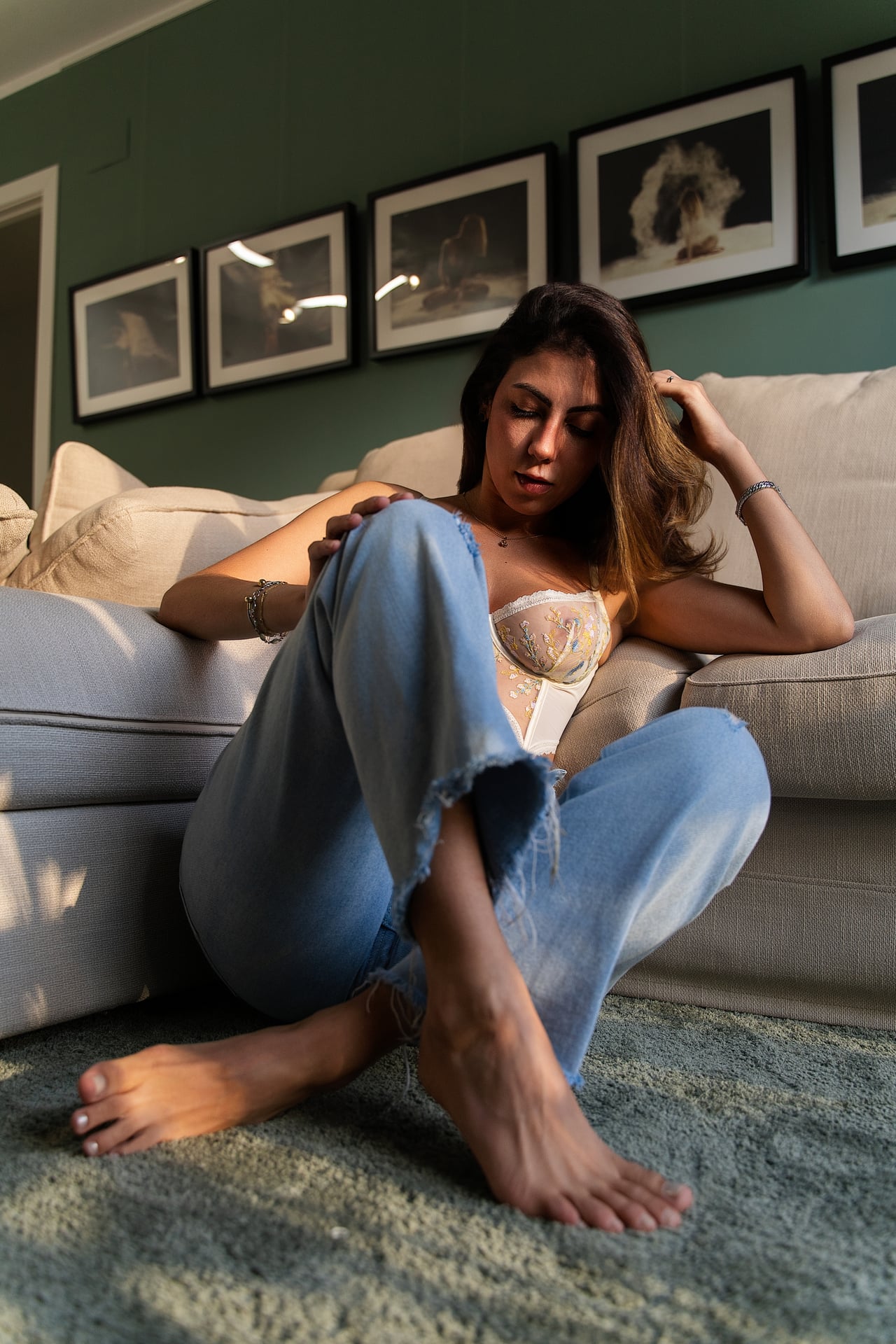 La Gabi seu a terra recolzada en un sofà beix, amb texans esquinçats blau clar i un bustier de lligenceria transparent, davant d'una paret verda amb fotografies emmarcades.