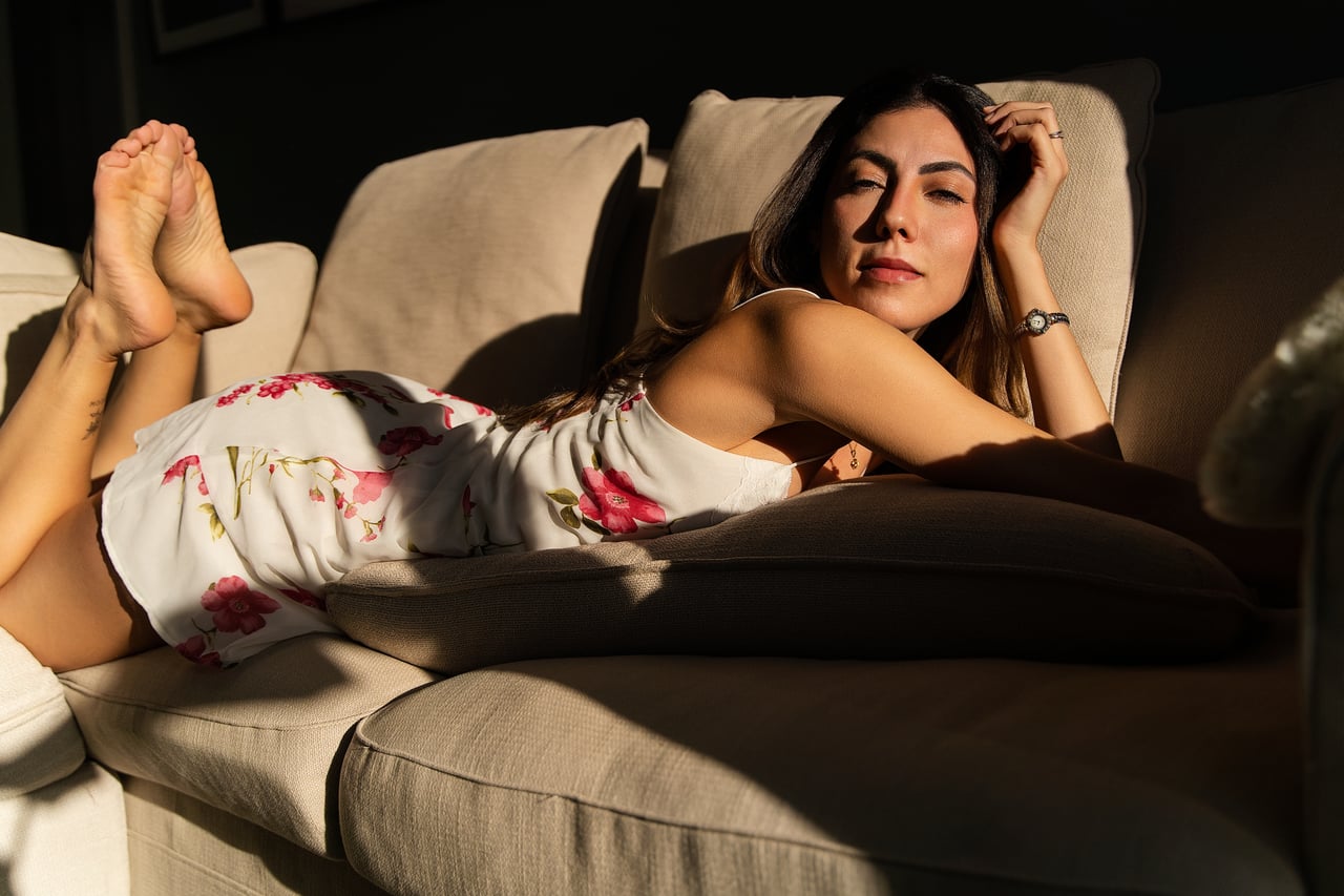 La Gabi, ajaguda de costat sobre un sofà beix amb els ulls tancats, porta un vestit blanc de flors i va descalça; la llum del sol li il·lumina la cara i el cos.