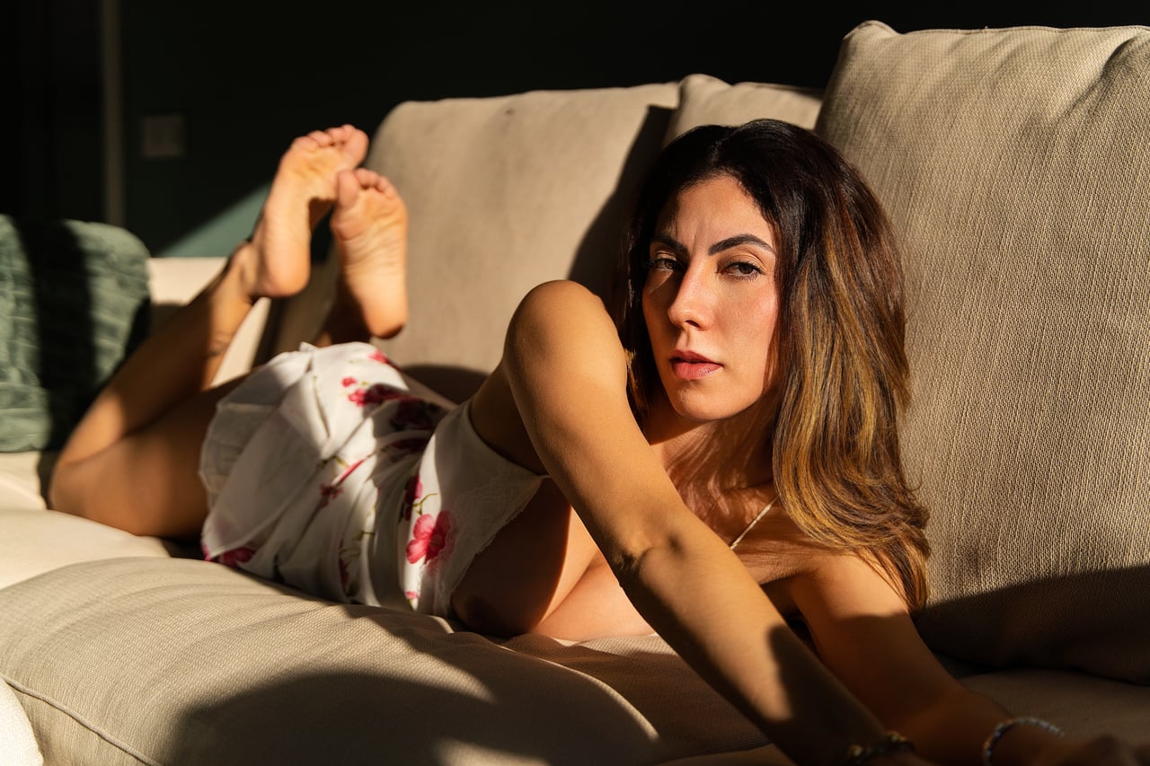 Dona ajaguda de costat sobre un sofà beix, amb el pit descobert i una peça de roba de flors sobre els malucs i els peus alçats, sota una llum solar marcada.