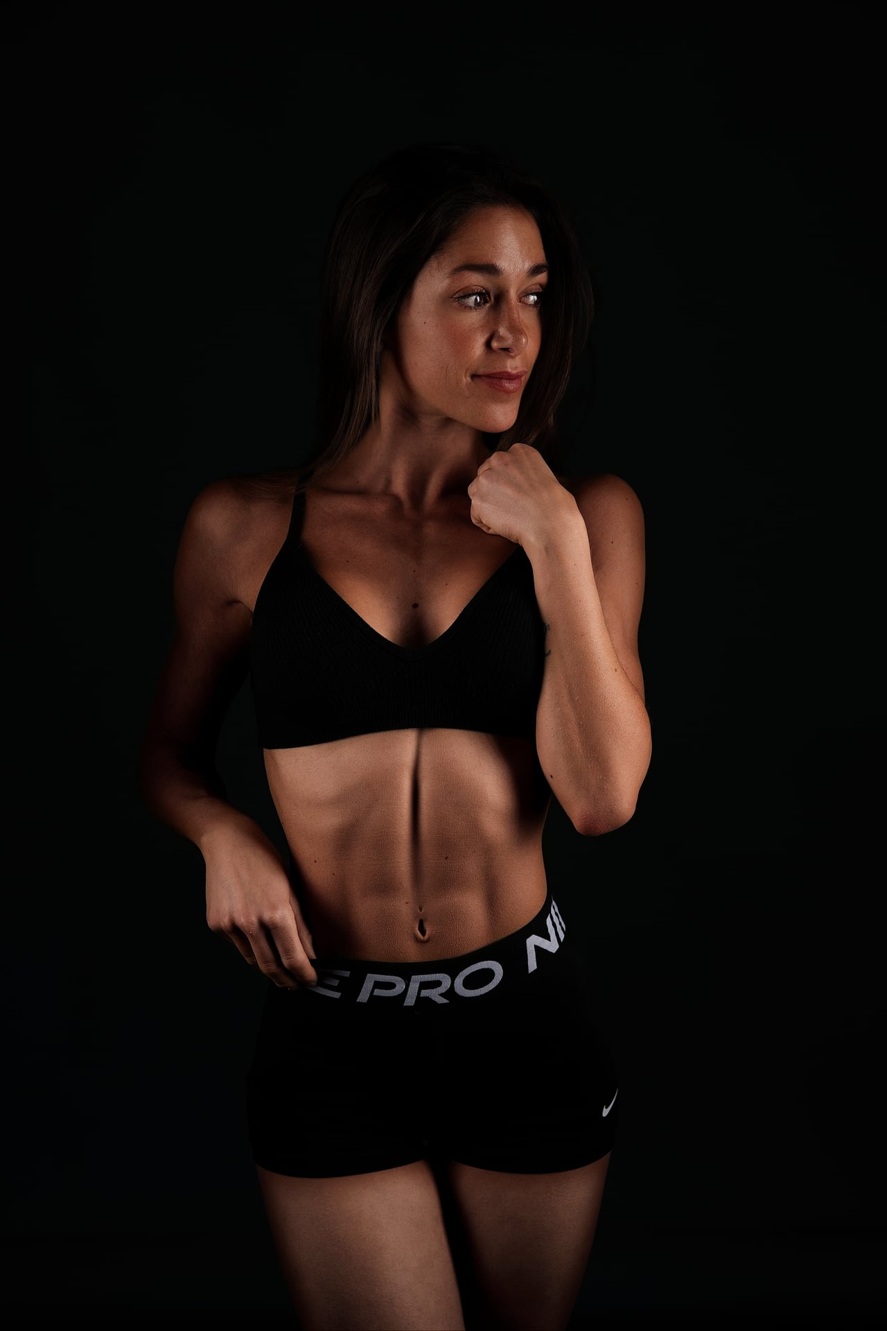 Dona amb top esportiu negre i pantalons curts Nike negres, dempeus amb el puny tancat a prop del coll i la mirada cap a un costat, sobre un fons d'estudi fosc.