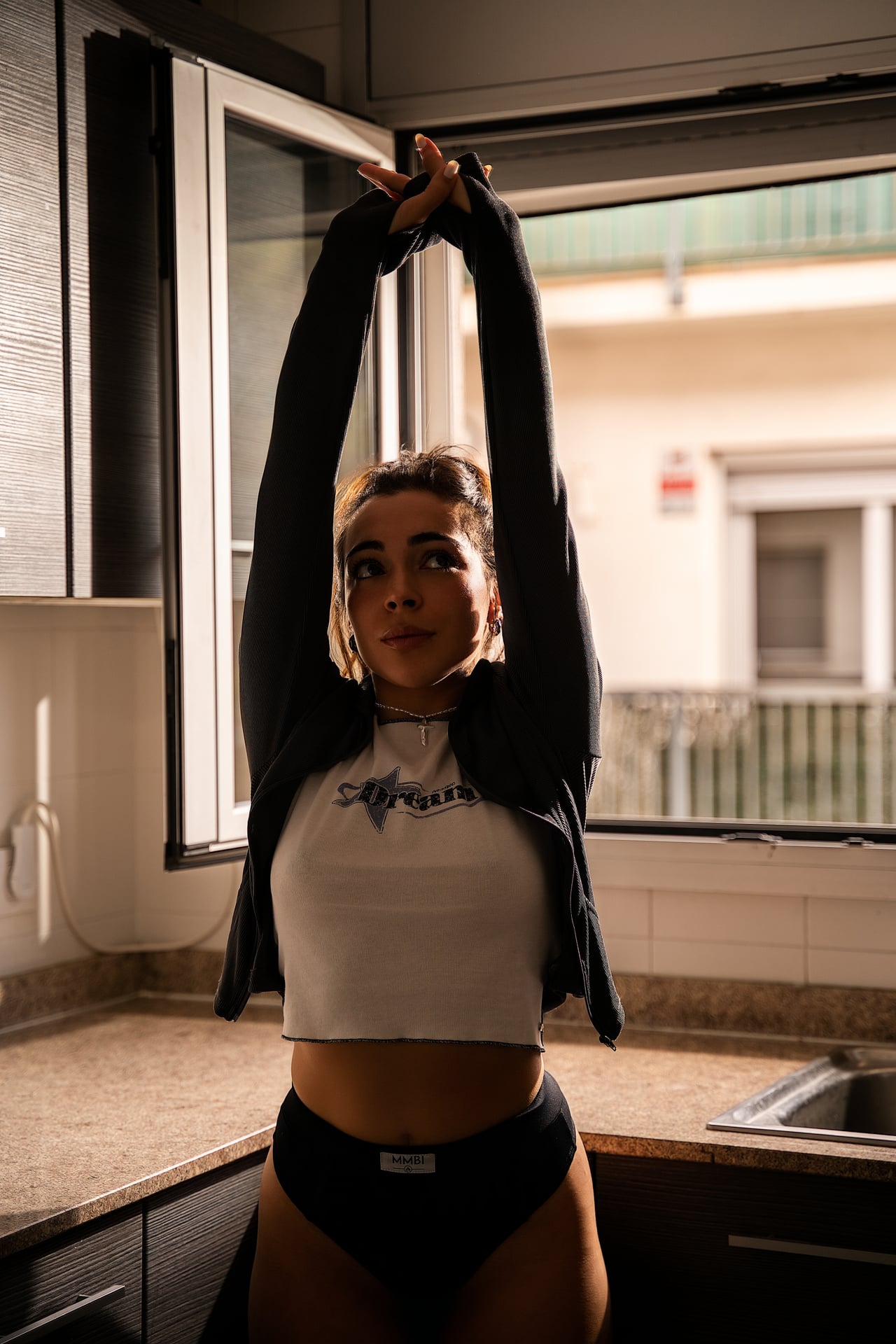 Una dona dreta a la cuina, amb els dos braços estirats cap amunt, vestida amb una samarreta blanca curta de màniga llarga i calces fosques, al costat d'una finestra oberta.