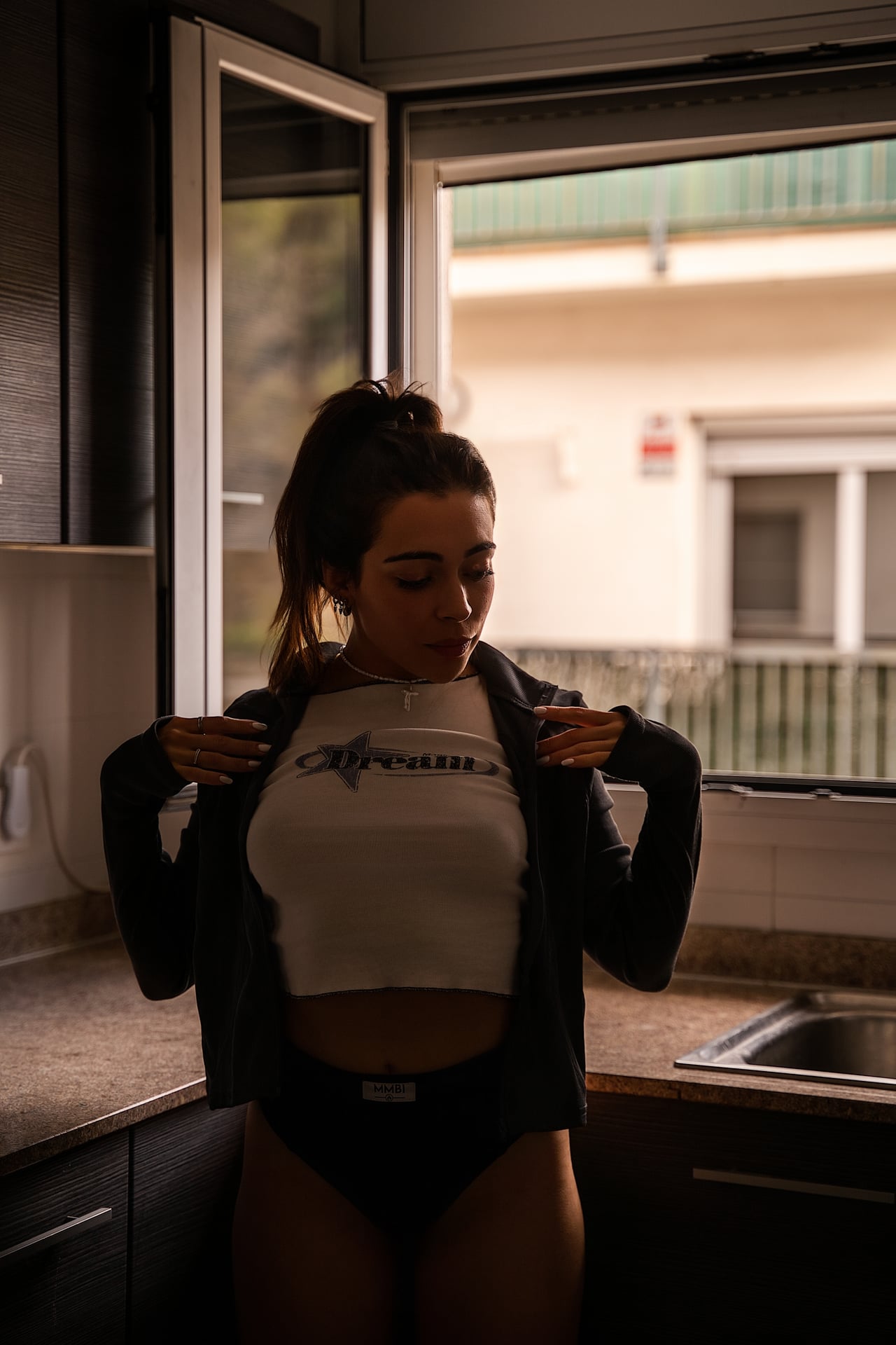 La Regina, dempeus a la cuina, s'aparta una jaqueta fosca oberta deixant veure un top curt amb l'estampat «Dream» i una calça fosca de cintura alta, amb el ventre i les cames descoberts, davant una finestra il·luminada.
