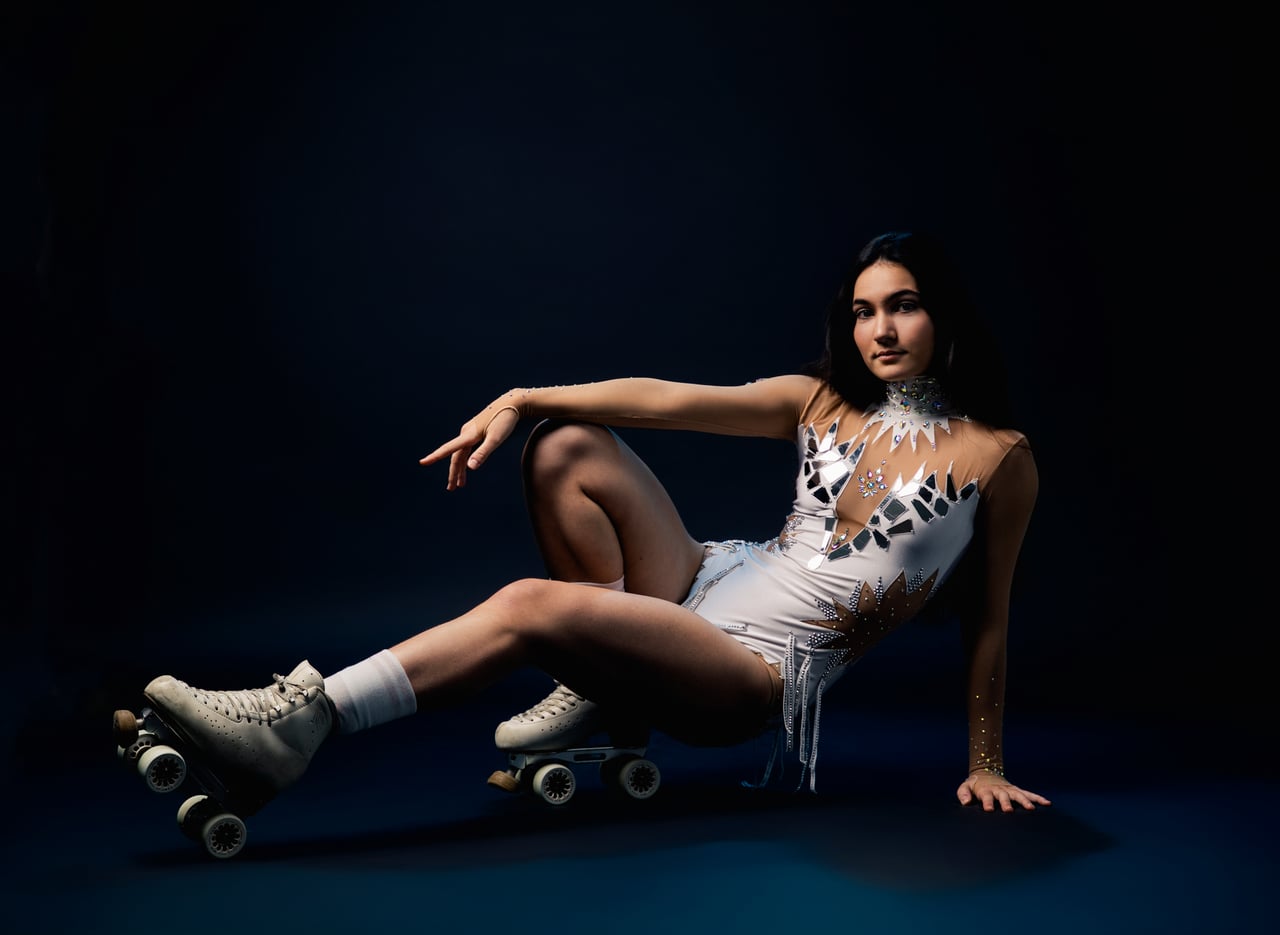 Dona asseguda a terra amb un vestit blanc de patinatge amb aplicacions platejades, mitjons blancs i patins de quatre rodes, sobre un fons fosc d'estudi.