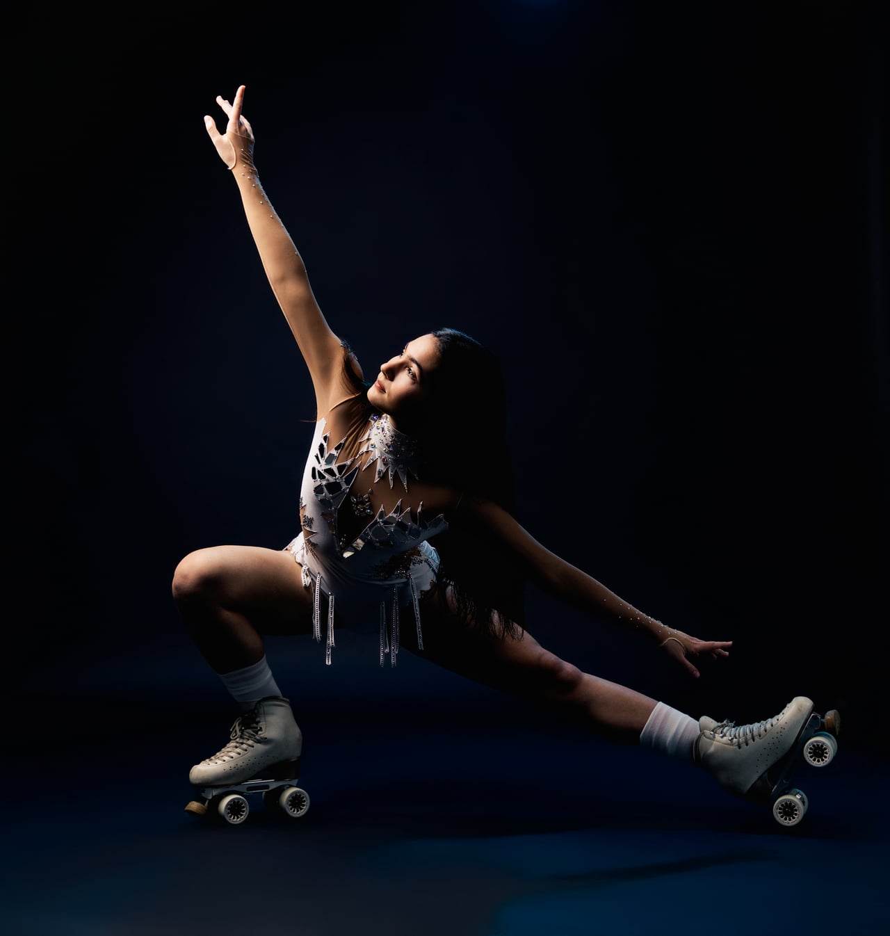 Dona en una posició de flexió baixa amb una cama estirada i un braç enlaire, vestint un mallot blanc brillant amb serrells, mitjons blancs i patins de quatre rodes, sobre un fons fosc d'estudi.