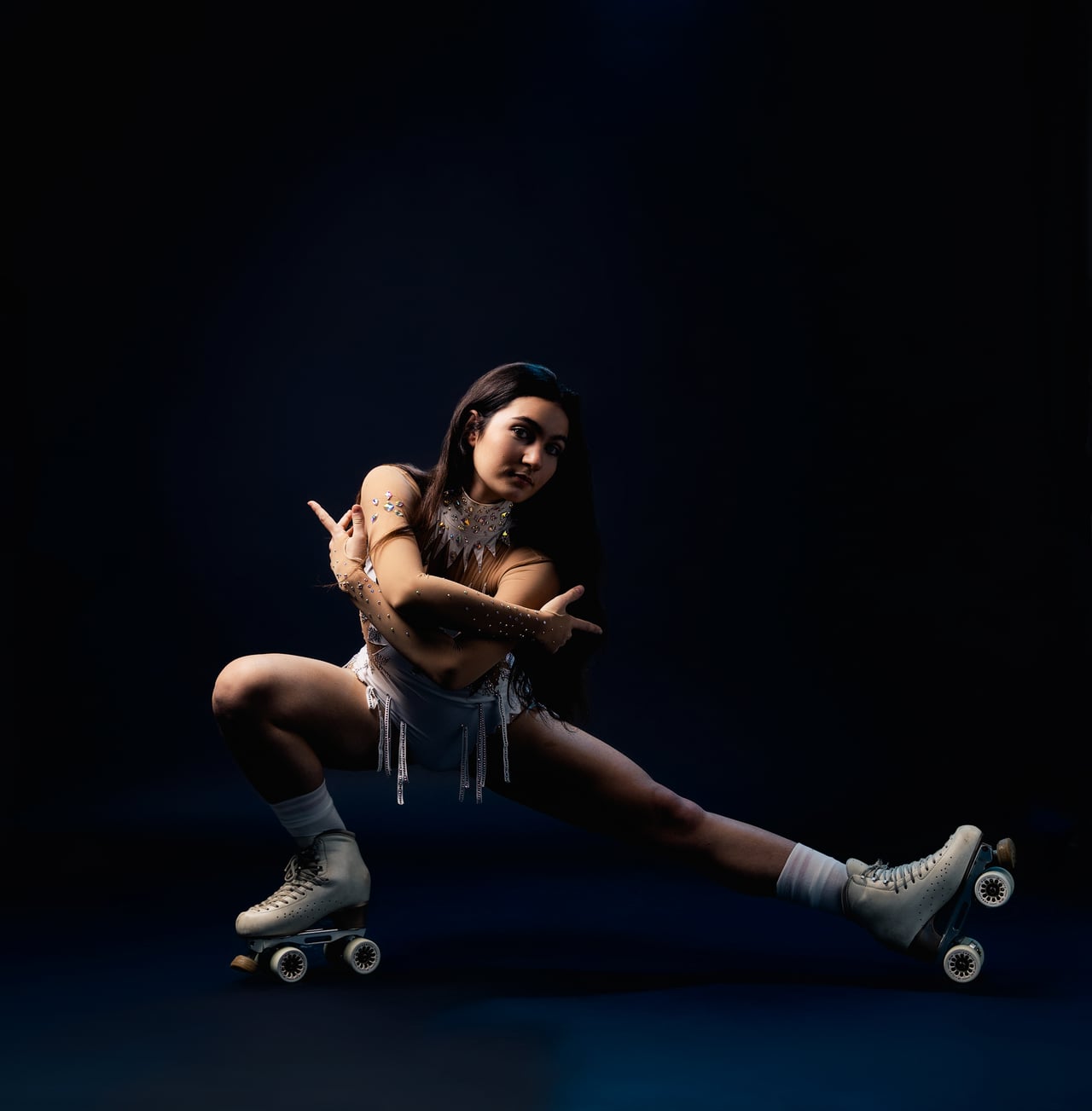 Dona amb un vestit de patinatge blanc brillant i patins de quatre rodes blancs, en una postura baixa d'equilibri amb una cama estirada i els braços creuats, sobre un fons d'estudi blau marí fosc.