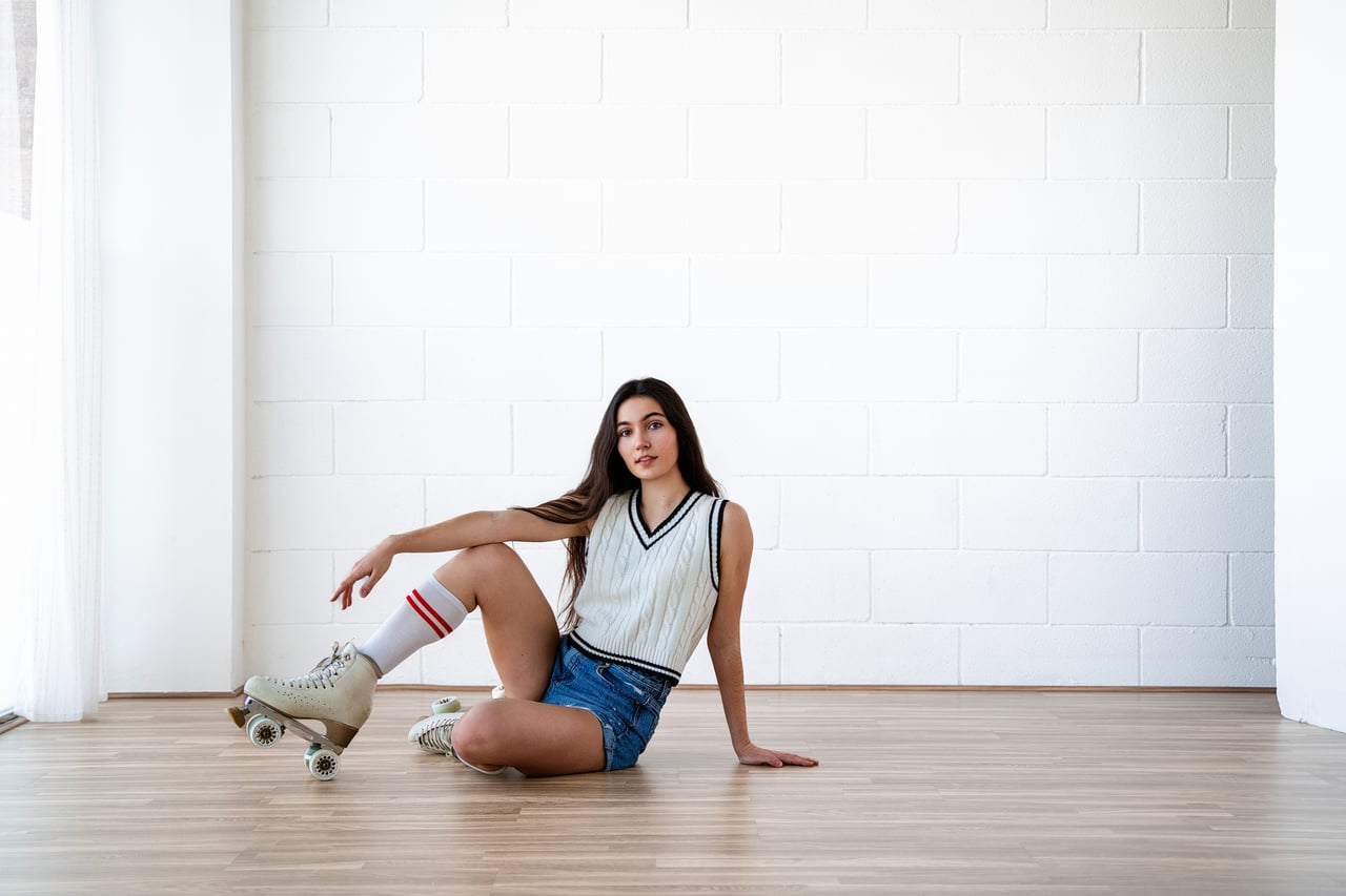 Dona jove asseguda al terra de fusta amb una cama flexionada, vestint jersei de punt sense mànigues, pantalons curts texans, mitjons alts i patins de quatre rodes, davant d'una paret de maó blanc.