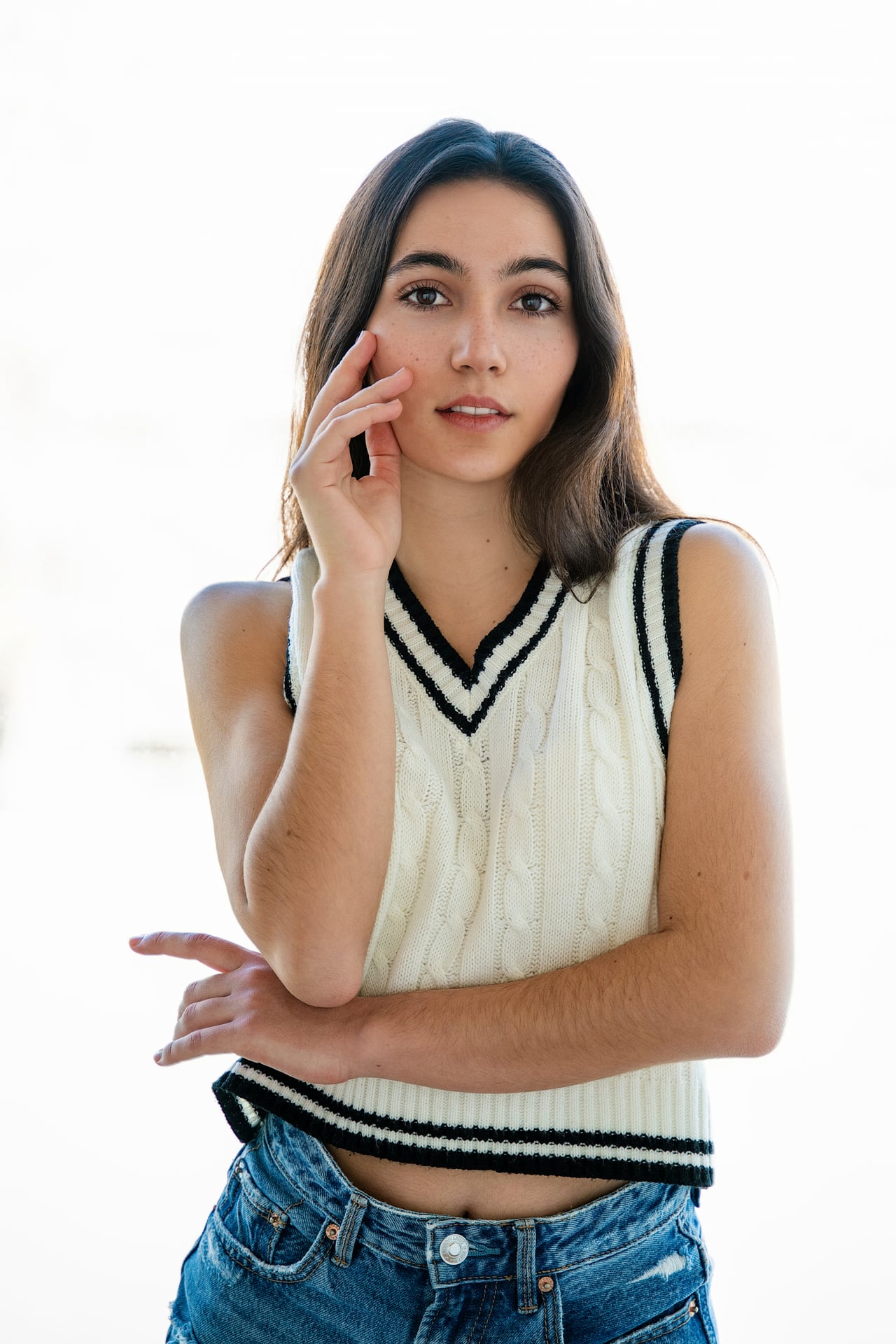 Una dona jove amb una armilla de punt blanca de coll en pic i pantalons curts texans es toca la galta amb una mà i creua els braços, davant d'un fons d'estudi blanc i molt il·luminat.
