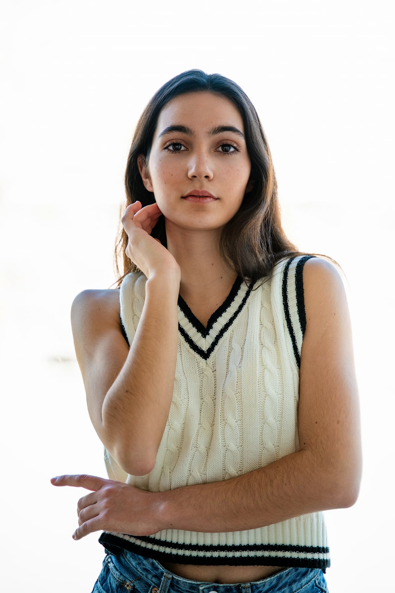 Dona jove amb cabell castany llarg, mirant a càmera amb la mà a la galta, vestida amb una armilla de punt color crema amb vores negres i pantalons curts de tela texana, en un estudi blanc molt il·luminat.