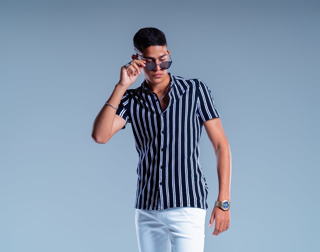En Simo, de mig cos, s'abaixa les ulleres de sol amb una mà; porta camisa de màniga curta de ratlles blanques i negres i pantalons blancs.