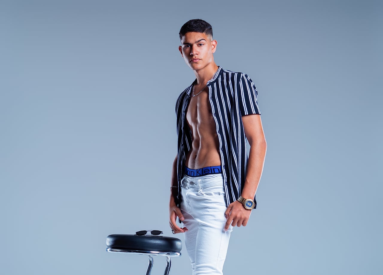 En Simo, de mig cos, dret amb la camisa de ratlles totalment oberta; s'intueix la cintura de la roba interior i porta pantalons blancs.