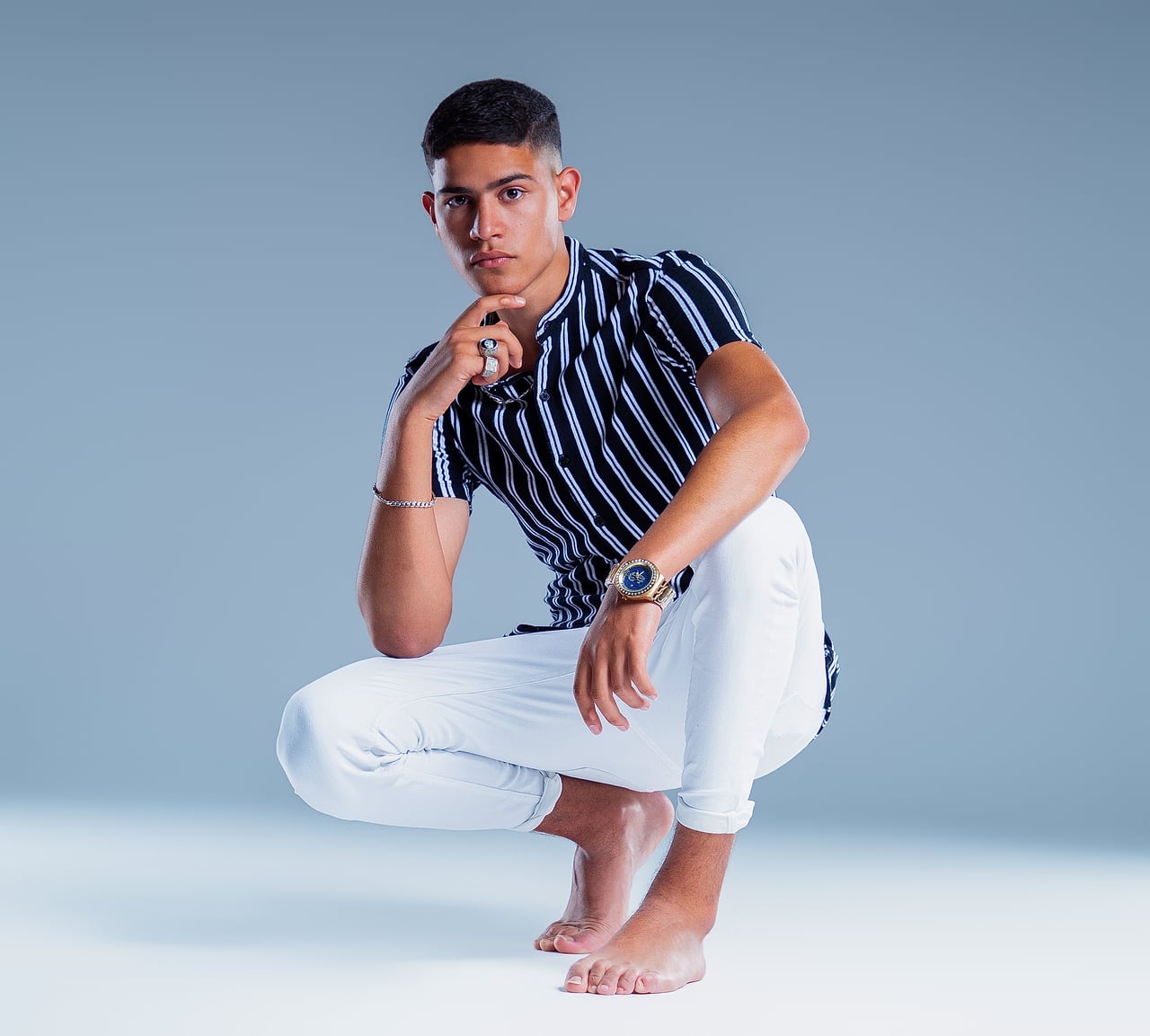 En Simo, de cos sencer, ajupit amb una mà recolzada a la barbeta; camisa de ratlles, pantalons blancs i descalç.