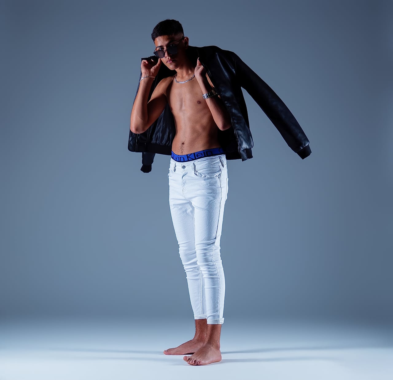En Simo, de cos sencer, dret amb una caçadora de cuir negre oberta sobre el tors nu; pantalons blancs i descalç.