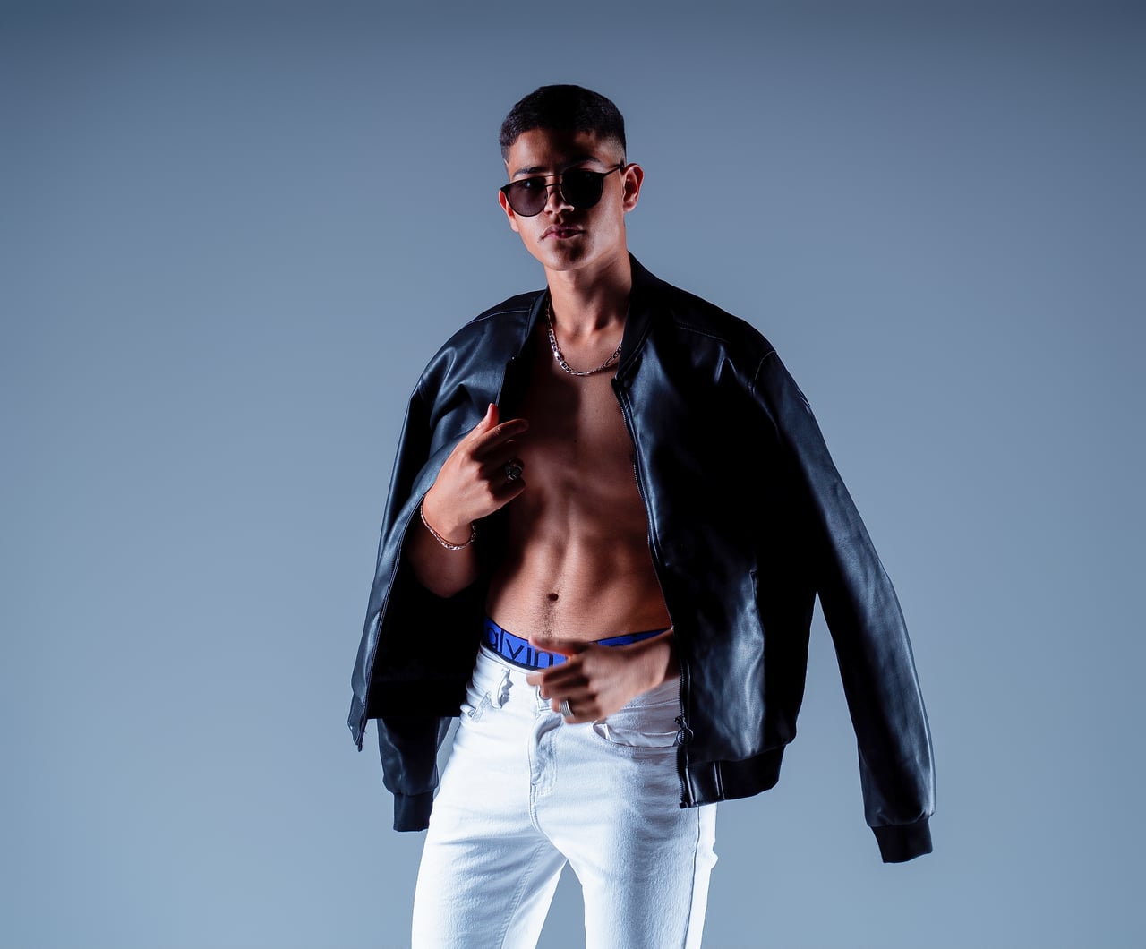 En Simo, de mig cos, amb una caçadora bomber de cuir oberta que deixa veure l'abdomen; ulleres de sol i pantalons blancs.
