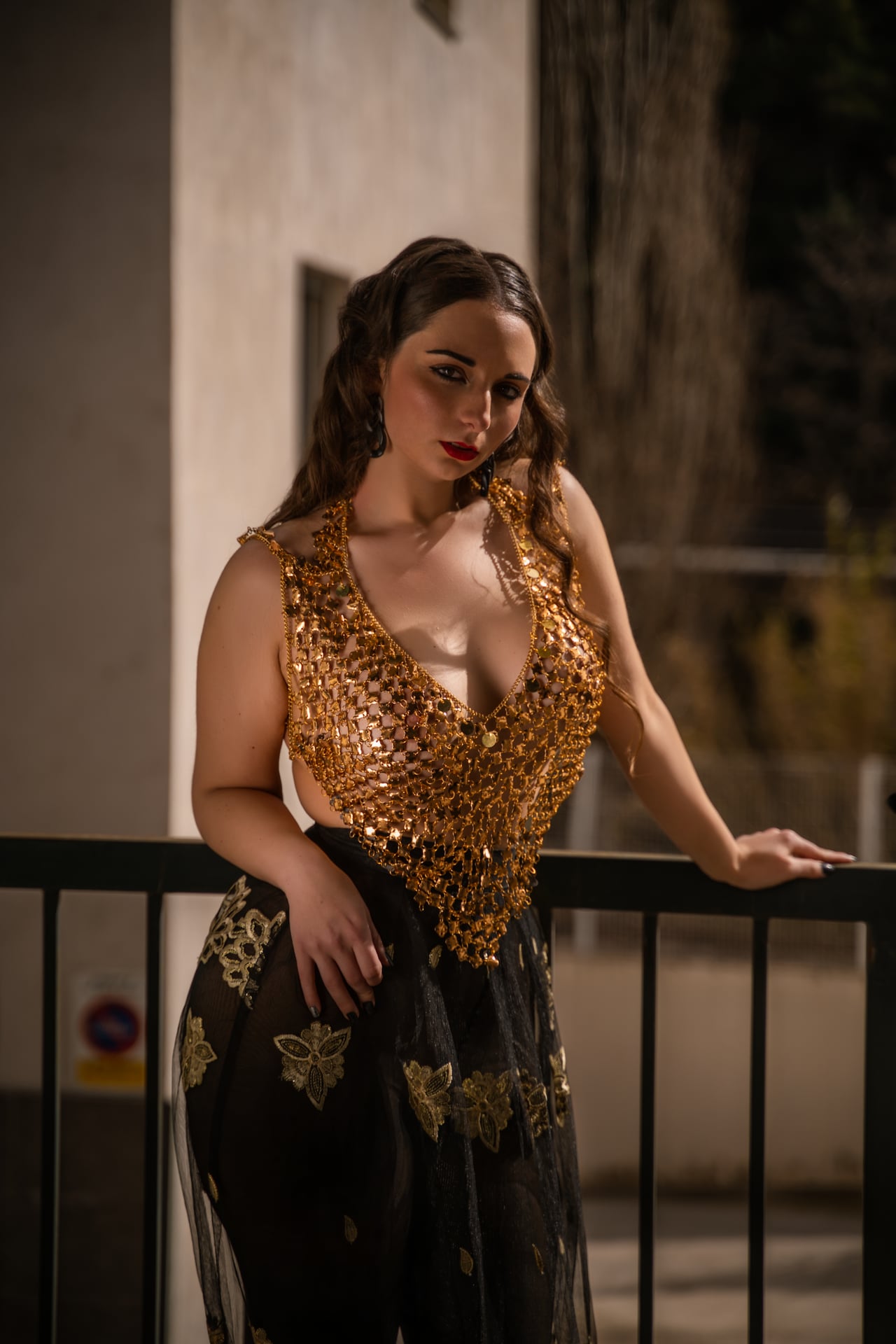 Dona dreta de nit recolzada en una barana metàl·lica, amb un top daurat de malla de lluentons amb escot en V profund i una faldilla negra transparent amb brodats florals daurats.