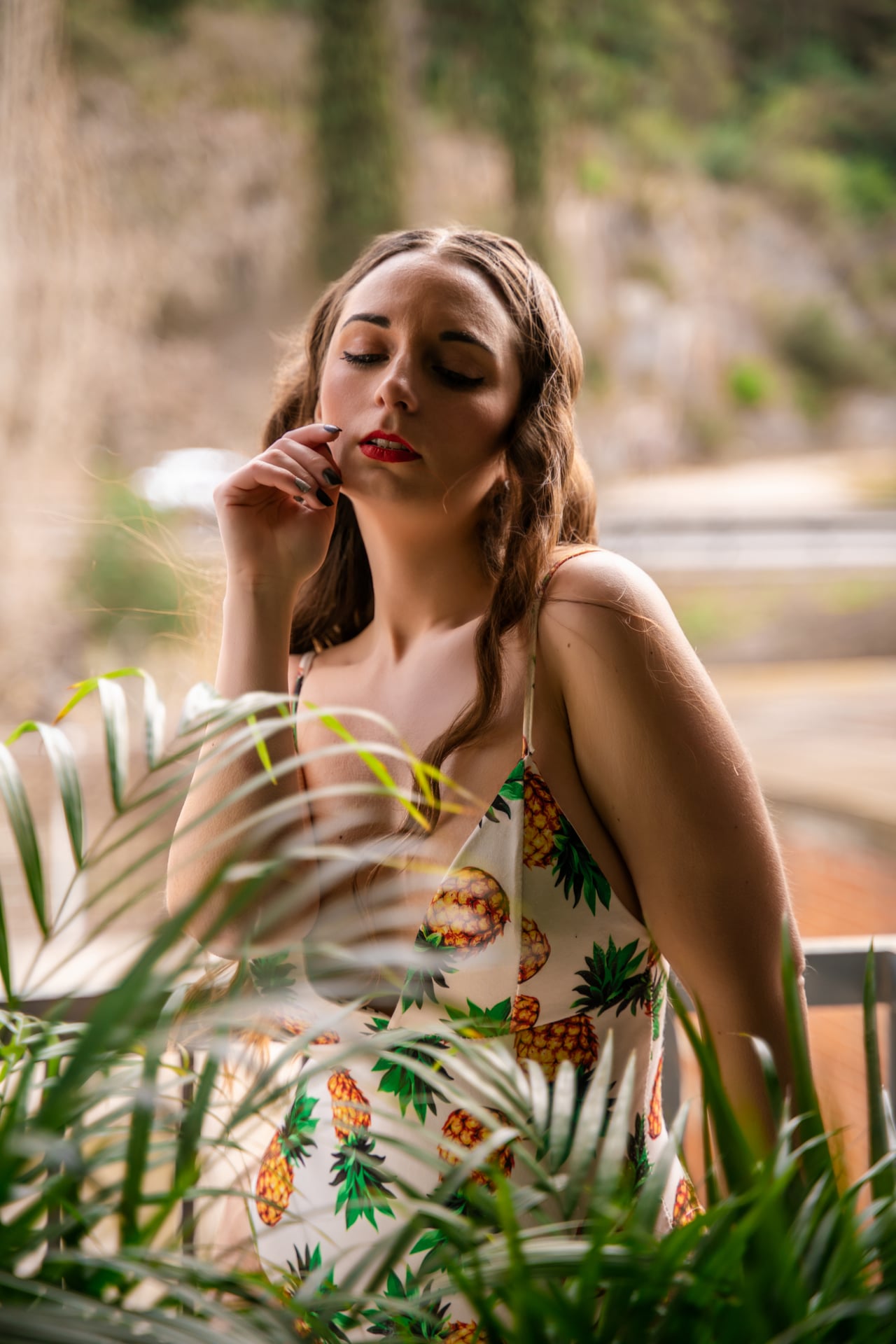 Dona jove asseguda amb un vestit blanc de tirants estampat de pinyes, els ulls tancats i la mà a la cara, entre fulles de palmera amb un fons exterior desenfocat.