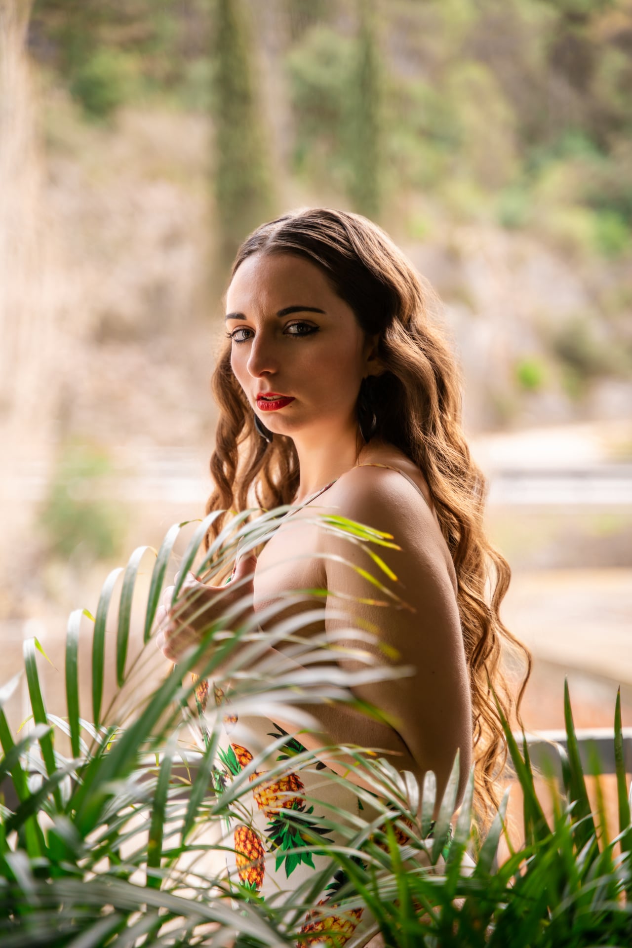 Dona jove de perfil i d'esquena, amb l'esquena i l'espatlla nues i un vestit estampat de pinyes lliscat avall, el braç li cobreix el pit, entre fulles de palmera amb llum natural càlida.